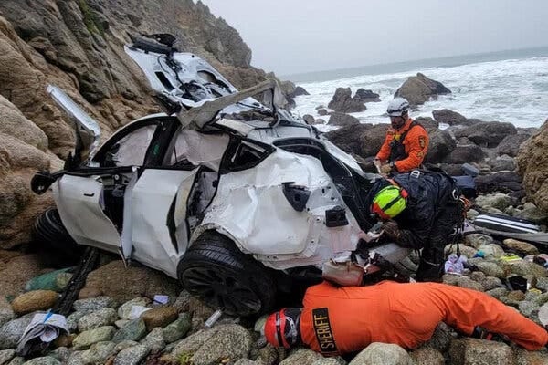 A white Tesla is severely damaged on a rocky beach. Rescue workers, some in orange jump suits, are surrounding it. 
