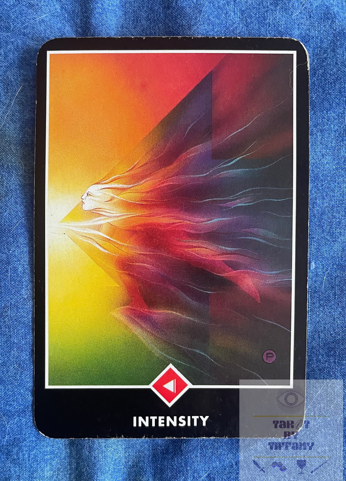 Image of the Knight of Wands (Intensity) card from the Osho Zen Tarot deck. The card is laid on a blue cloth background. The cad shows a person in a red, yellow, and purplish whirlwind that comes to a point like an arrow facing left. It is a dynamic image that invokes fast movement.