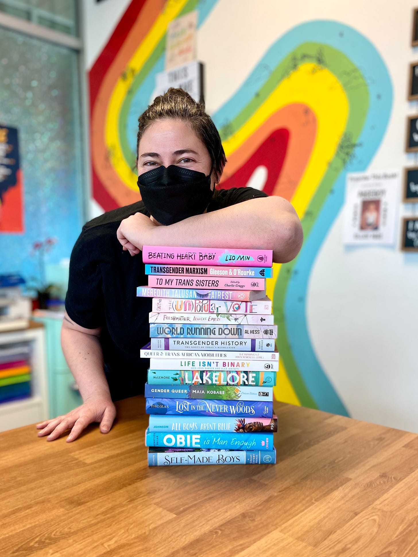 Kaitlyn, bookstore owner and white agender person, leans over a tall stack of queer books on a wood table. They are smiling behind a black mask and wearing a black t-shirt. In the background is a rainbow painted wall.