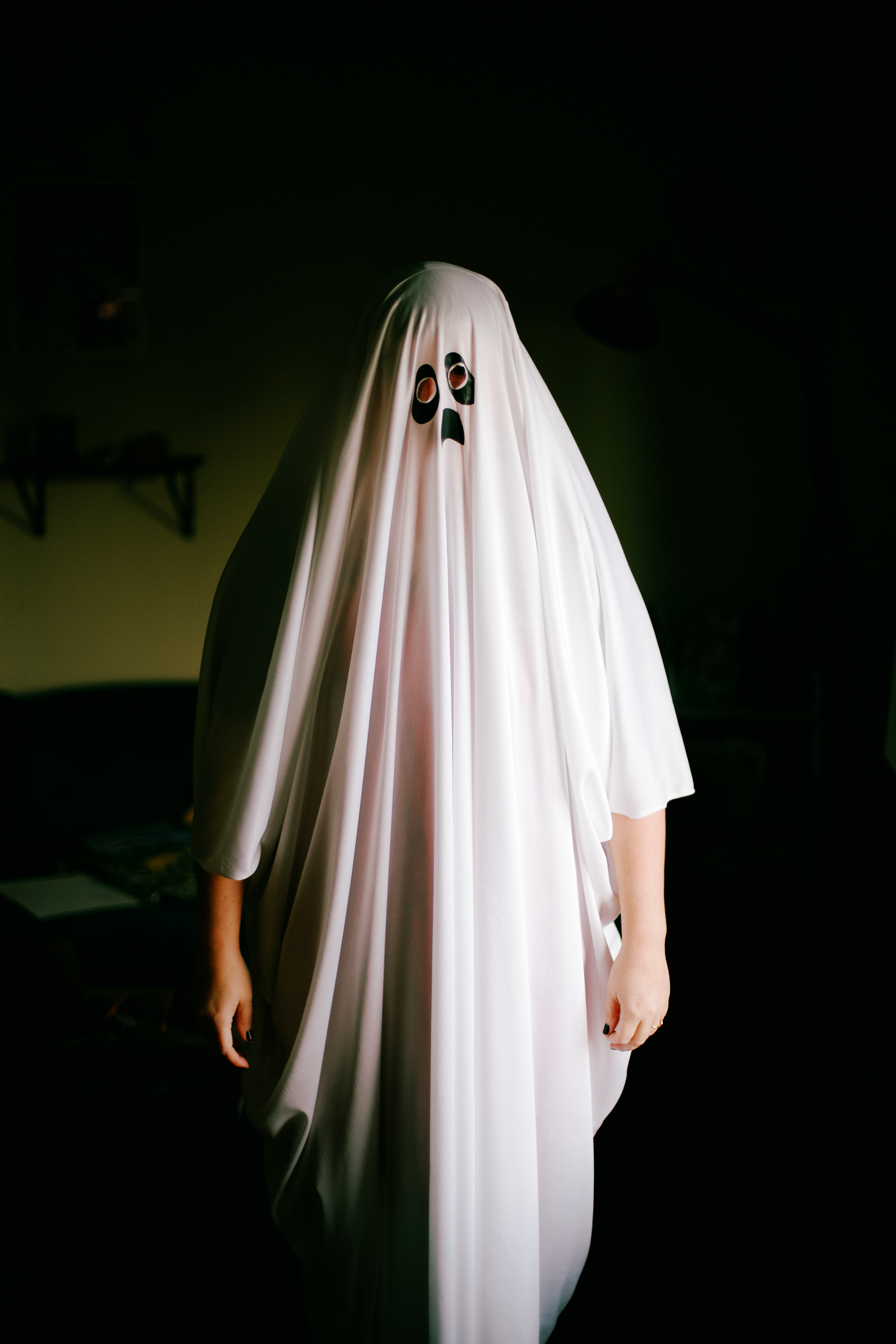 the background is a moody black and olive green. A person standing under a white sheet with two black eyes and a sad mouth 