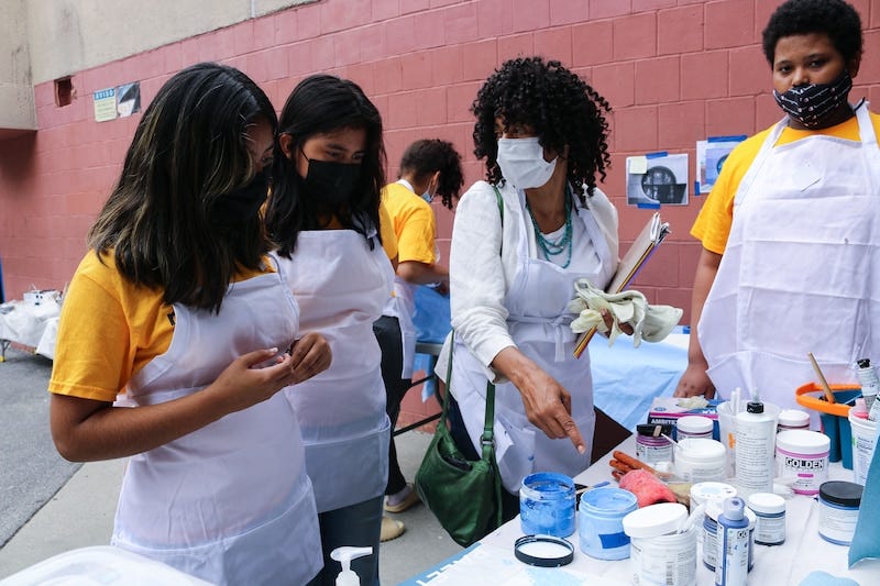 Four students wearing masks outside and aprons work outside on a table full of paints.