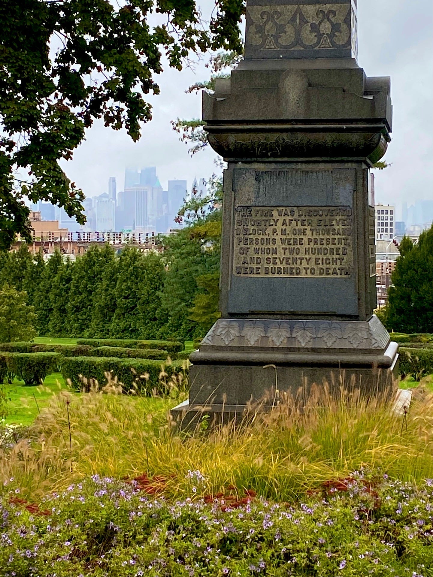 The memorial for the Brooklyn Theatre fire and the marker for its unknown victims. A bronze plaque on an obelisk set amongst grass and flowers reads, "The fire was discovered shortly after 11 o'clock, P.M. One thousand persons were present of whom two hundred and seventy eight were burned to death." The skyline can be seen in far background.
