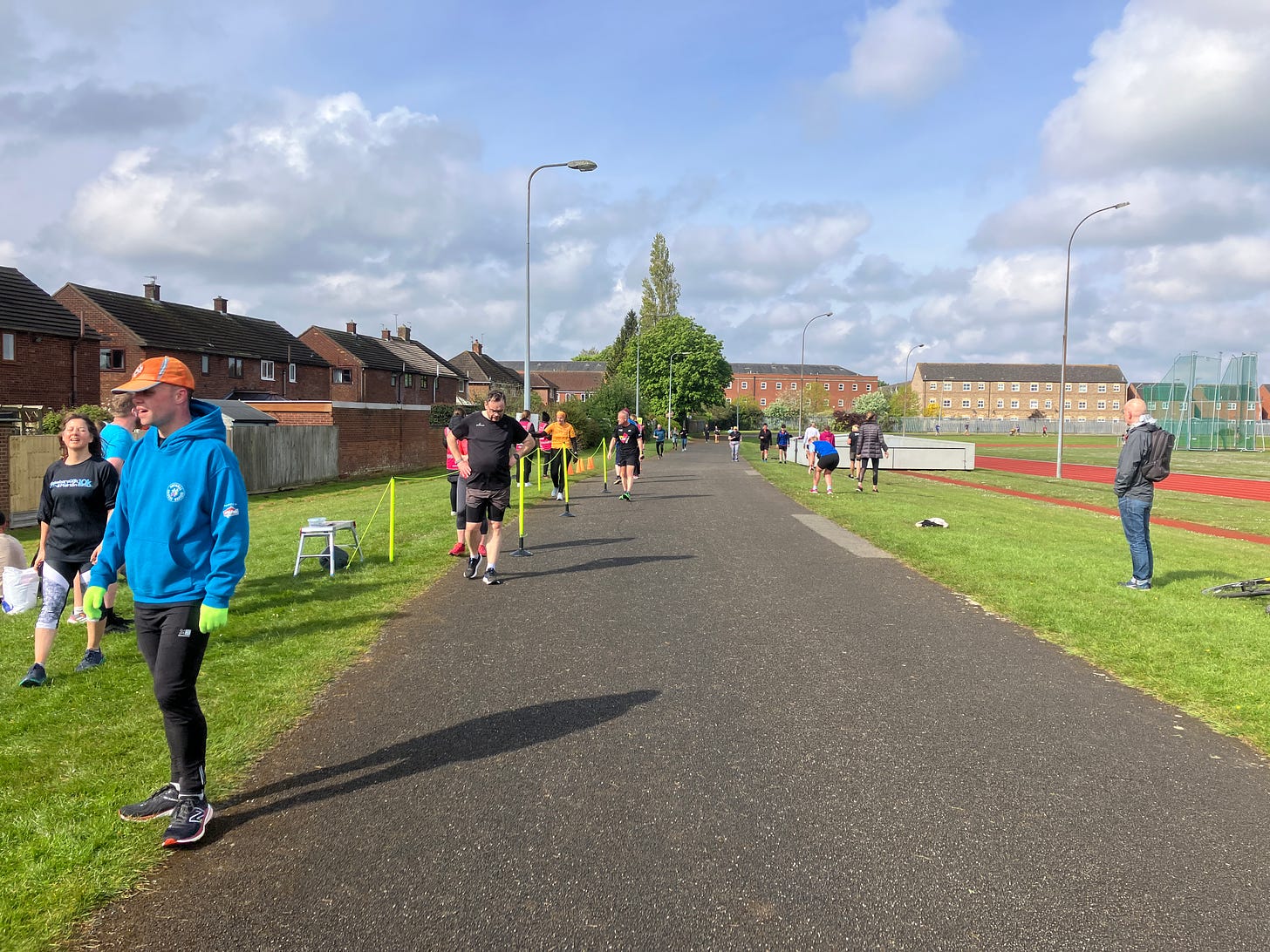 Participants walking and jogging towards the finish line on a sunny day, with onlookers and a backdrop of a track and residential buildings