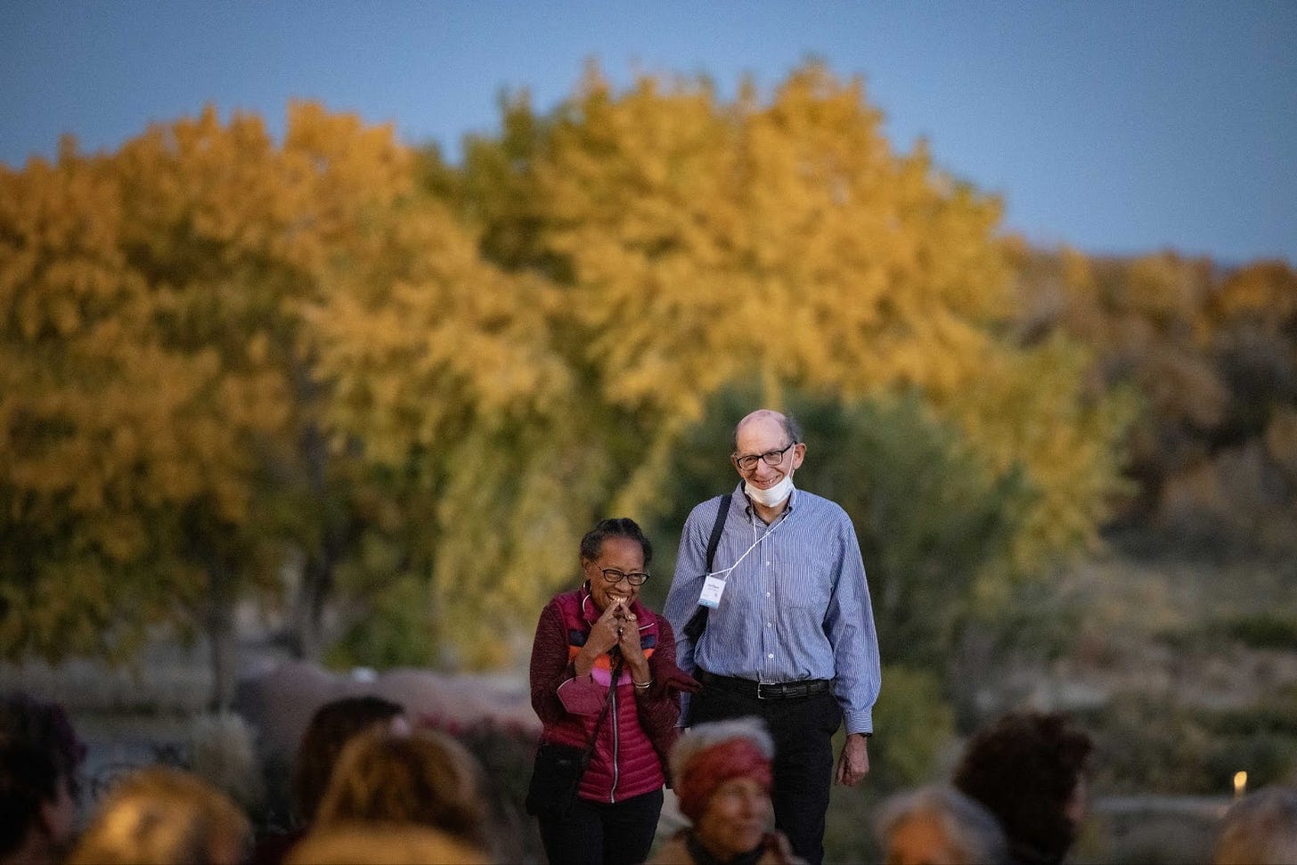 A picture of a tall, older white man in a button down shirt walking next to a shorter, older, black women. Both are smiling. It is dusk with a beautiful stand of trees behind them.