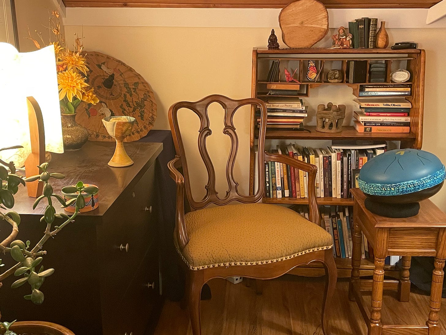 An empty wooden chair set in front of a bookshelf with a table lamp on one side and a blue steel tongue drum on the other side.