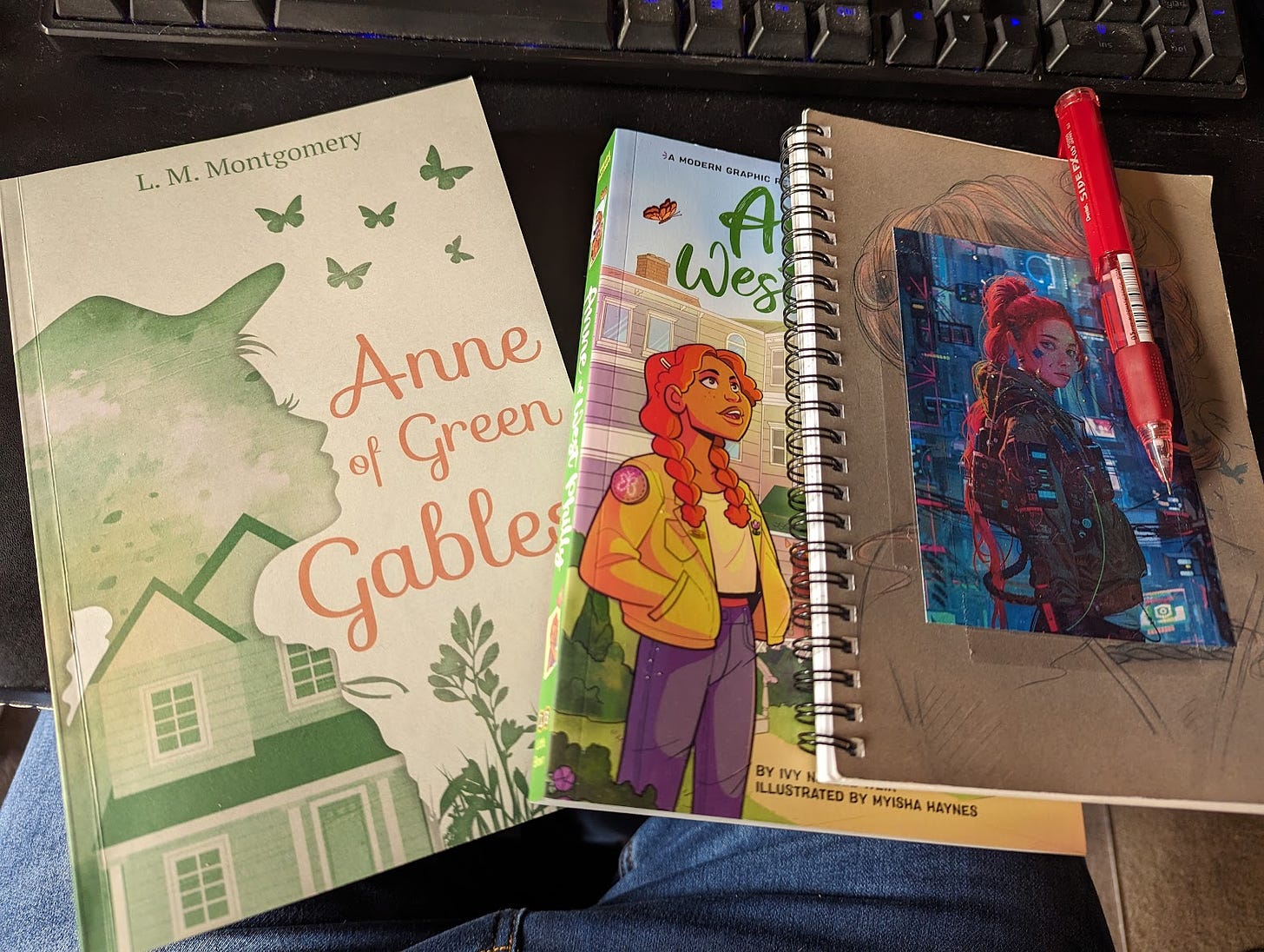 Three books on a computer table. "Anne of Green Gables" by L.M. Montgomery, "Anne of West Philly" by Ivy Noelle Weir, and a notebook with a cyberpunk Anne Shirley stickytaped on the front.