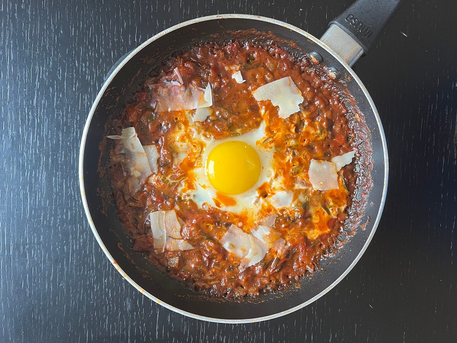 A rich and flavorful shakshuka with a deeply cooked tomato base, topped with a runny egg yolk and thin shavings of Parmesan. The sauce has a deep, almost caramelized color, showing the intensity of the slow-cooked ingredients. This is one of Liat Portal’s homemade shakshuka creations, part of her Foodie Disorder journey, where she explores the layers of flavor in simple yet complex dishes.