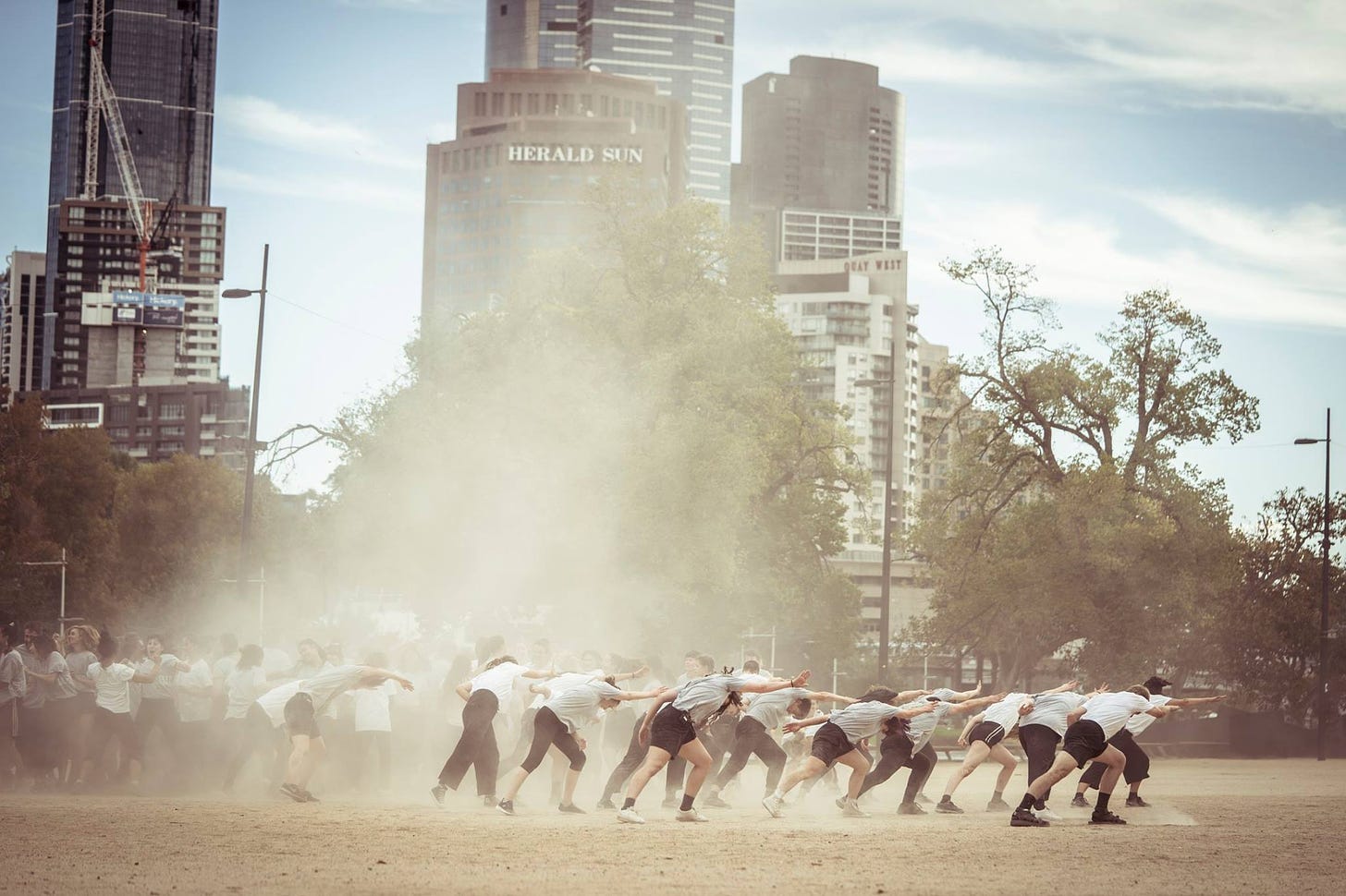 One group of dancers leans to their left, the other group rvaes, face-to-face. Dust rises against a backdrop of Melbourne's city buildings