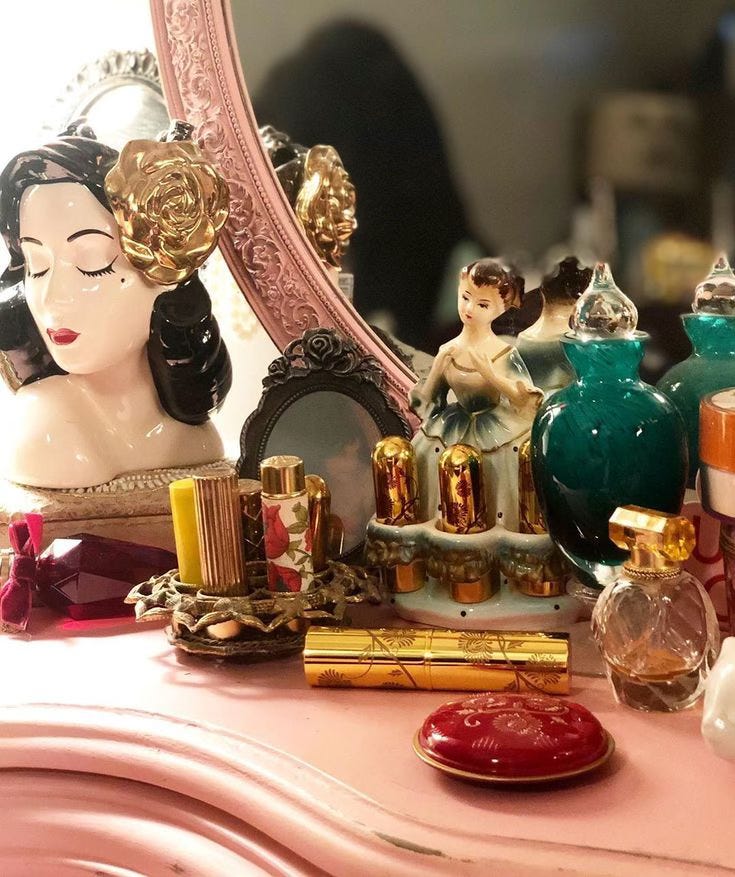 A vintage vanity setup featuring a porcelain bust with a golden rose hair accessory, ornate perfume bottles, gold and floral lipstick tubes, and a ballerina figurine.