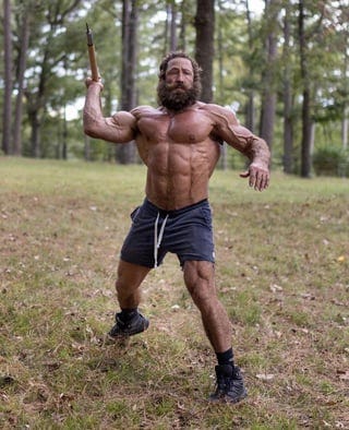An image of Liver King, a fitness influencer who falsely claimed not to use steroids. He stands in shorts with his chest bared, holding a wooden spear.