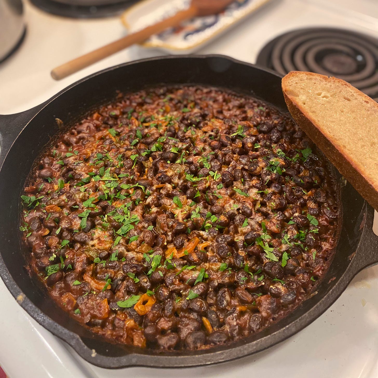 A cast iron filled with black beans baked in a tomatoey sauce with cheese and parsley on top. A slice of sourdough rests at the edge of the pan.