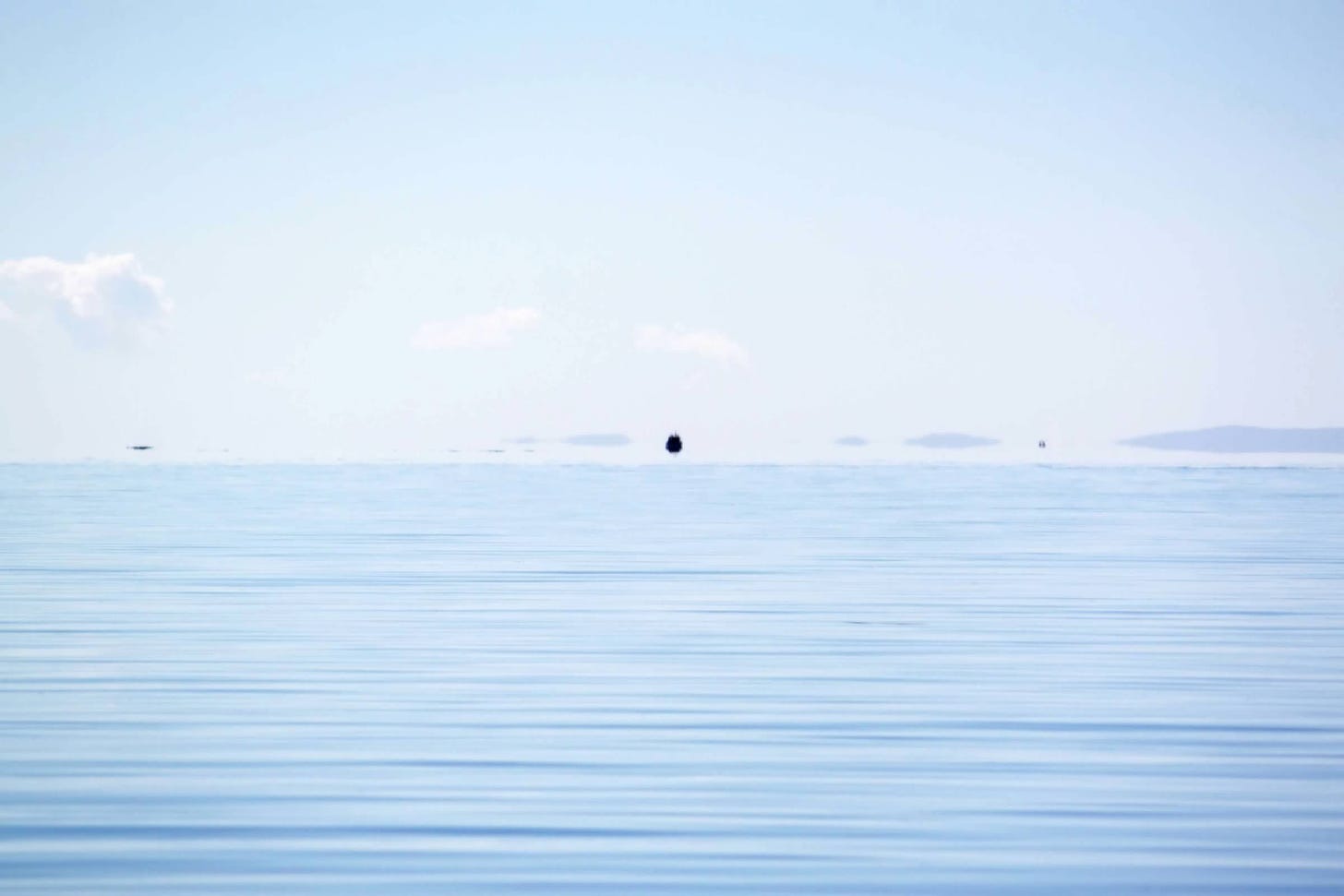 A calm blue lake with clouds on the horizon and a black dot (a tiny island) in the distance