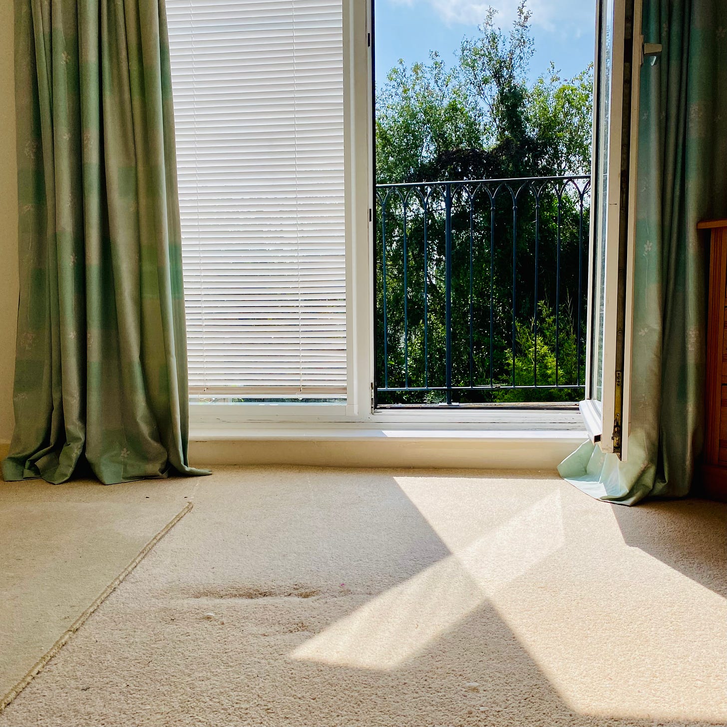 an empty room with french windows framed by blue curtains. One window is open onto a balcony with sunny blue sky, clouds, tree tops. The light from the sun casts a golden shadow into the room. 