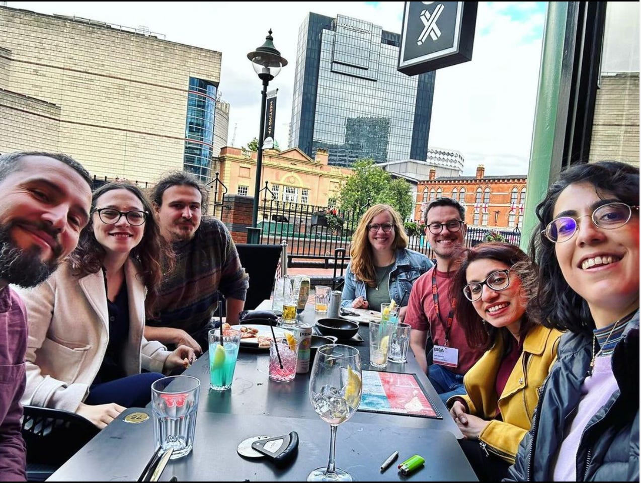 Six smiling people sitting around a long table outside with cocktails