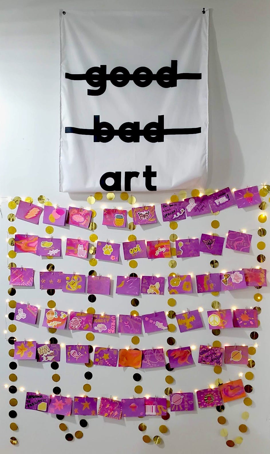 A white flag with black text reading "good (crossed out) bad (crossed out) art" hangs above a collection of purple and postcards, decorated by Uproar participants. They are hanging among large gold sequins on strings of twinkle lights.