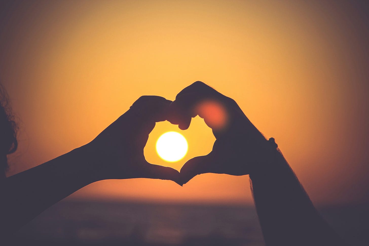 A photograph of two hands silhouetted against the setting Sun. The hands are joined together to form a heart shape.