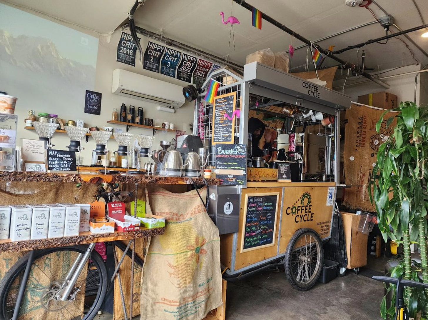 A profile of a coffee cart built on a bicycle frame that has been rolled into a retail space. A coffee bar made of live edge wood has been been built up over the bicycle's front tire and is covered in hot water kettles and pour over devices. A lower shelf holds tall white tins filled with coffee beans for sale and burlap coffee sacks hang underneath the shelves covering up the guts of the machine. A video screen projection of a mountain is on the back wall under a pink flamingo hanging next to a mini-rainbow flag from a water pipe near the ceiling.