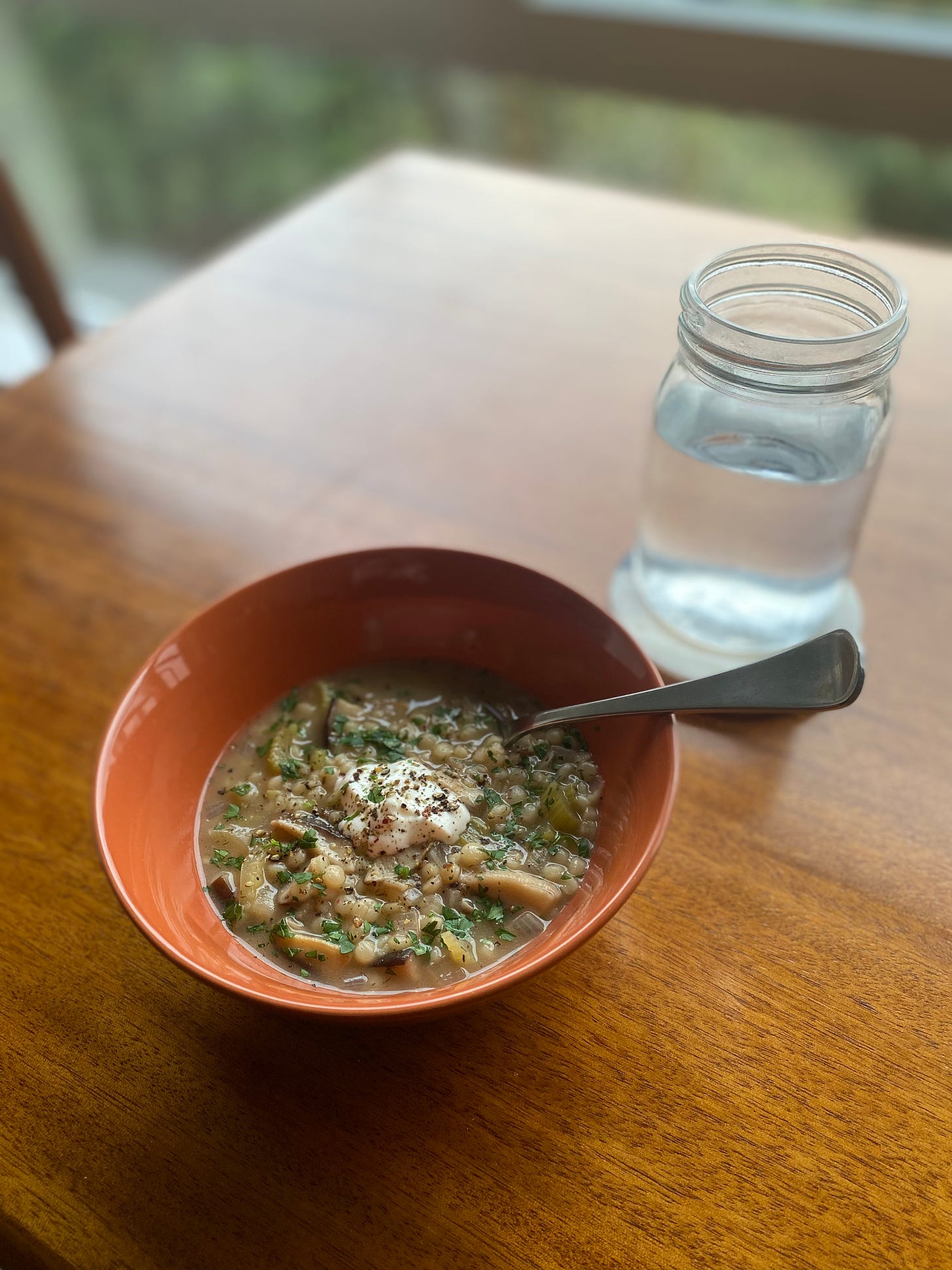 An orange bowl of the mushroom and couscous soup described above, dotted with parsley and black pepper, with a dollop of sour cream in the middle. A spoon sticks out of the bowl on the right side, and a jar of water is on a coaster next to the bowl on the table.