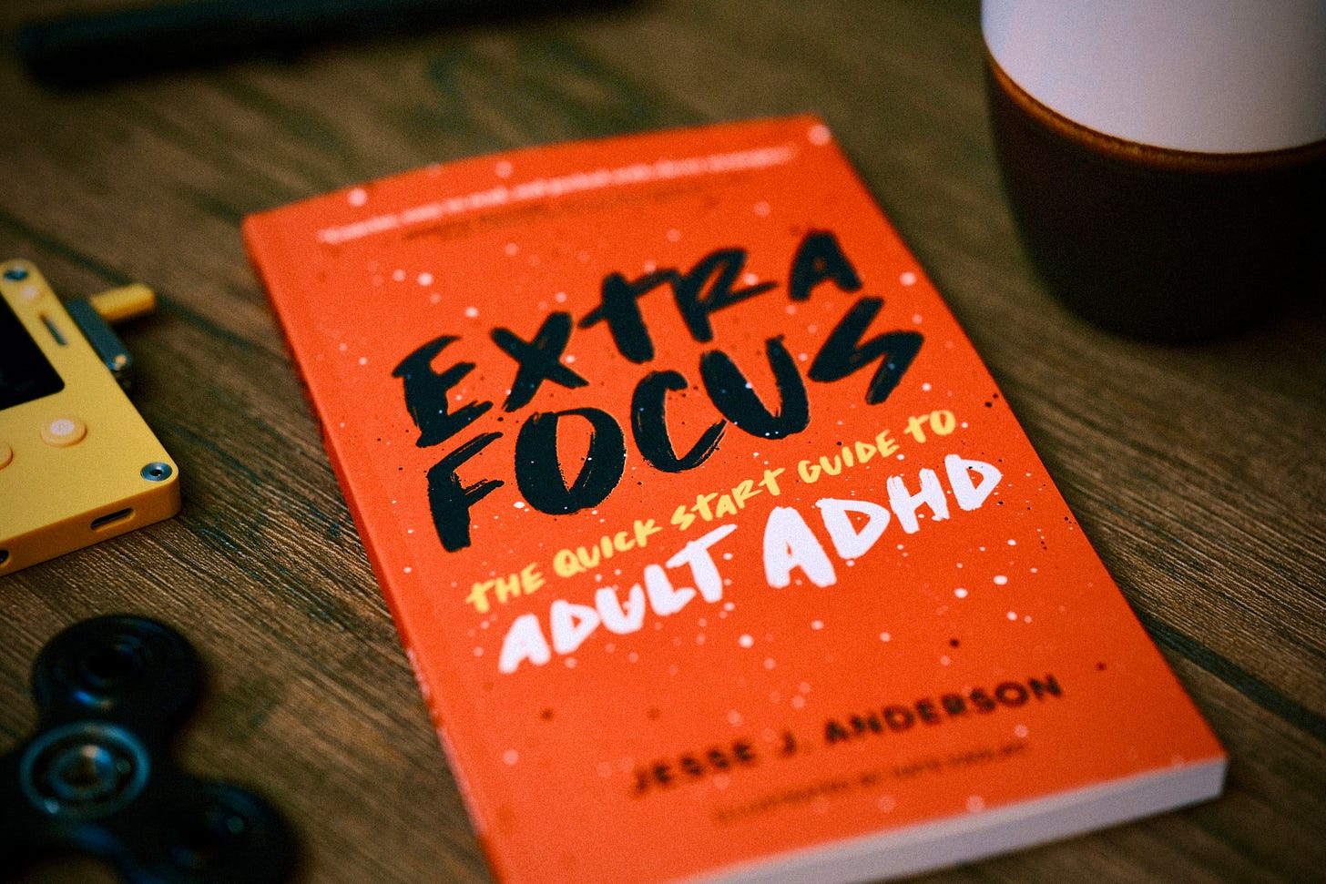 An artistic photo of the Extra Focus: The Quick Start Guide to Adult ADHD book, on a wooden table surrounded by some figet toys and a cup of coffee. On a lovely dark wooden table.