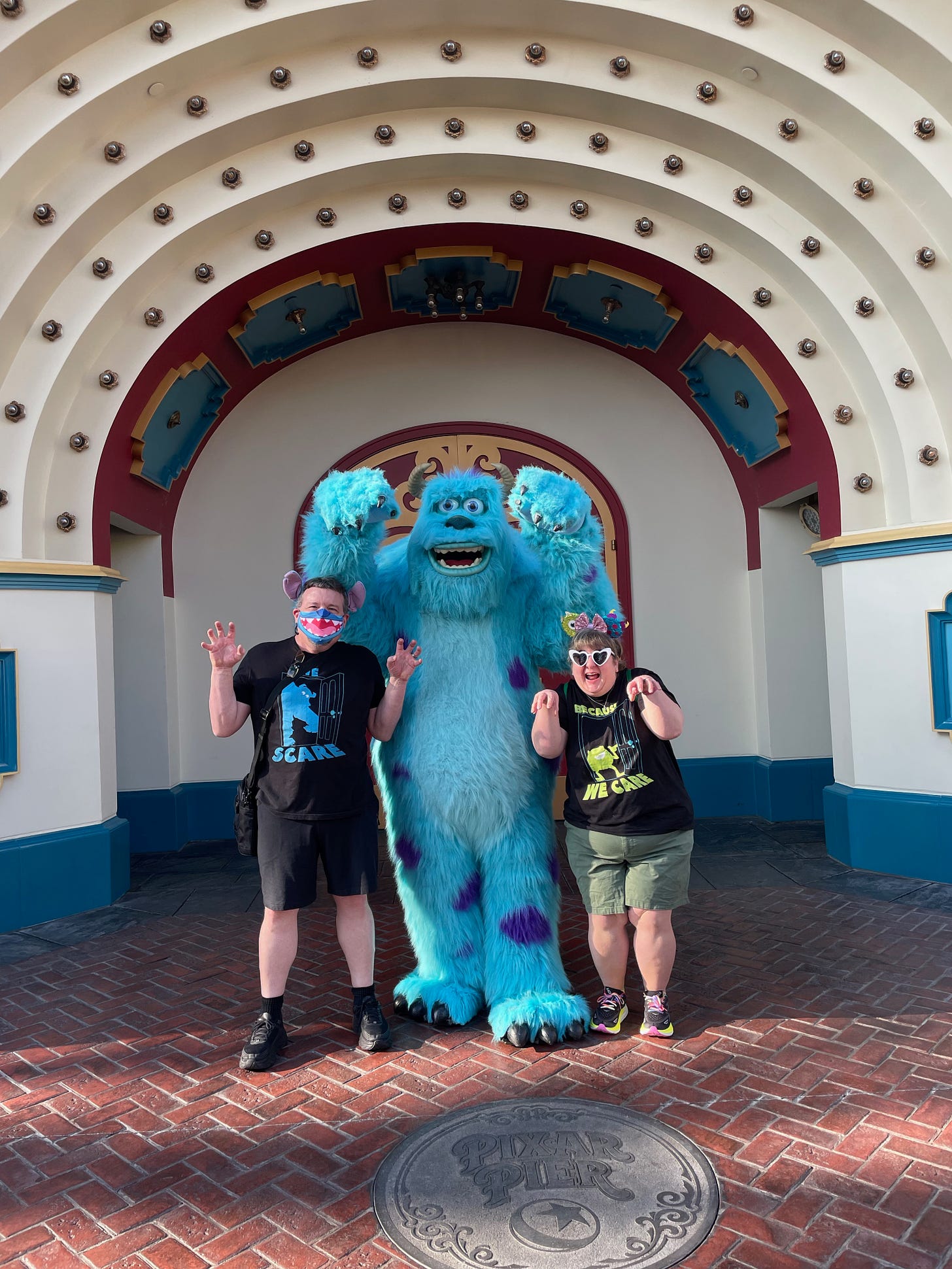 Two grown ass adults wearing ear headbands and coordinating Monsters, Inc shirts, in a "scare" pose with Sully.
