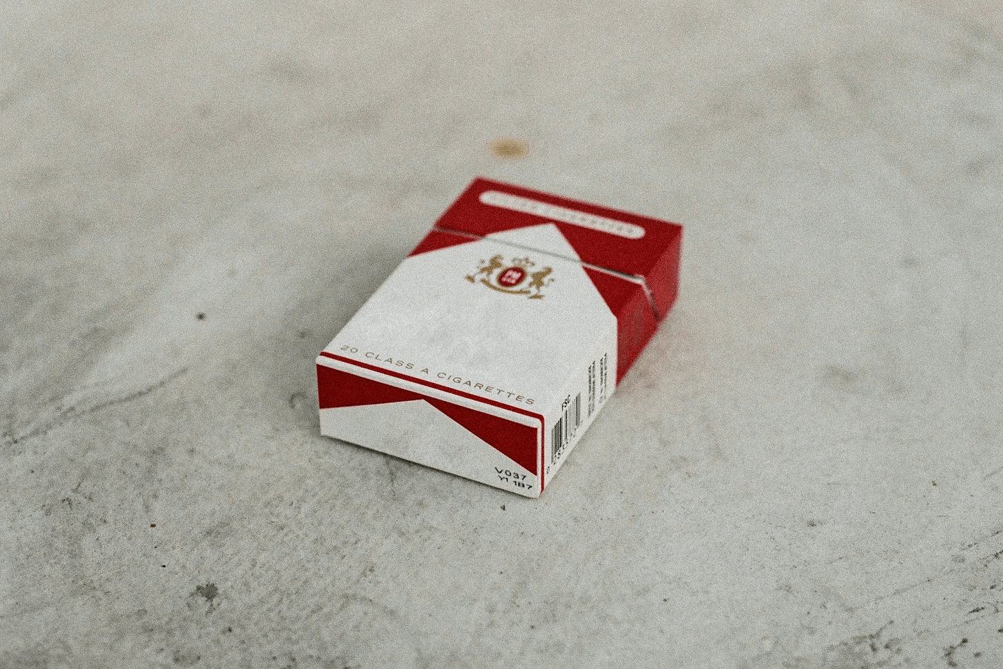 Uma caixa de cigarro branca e vermelha apoiada em um chão de cimento cinza claro.