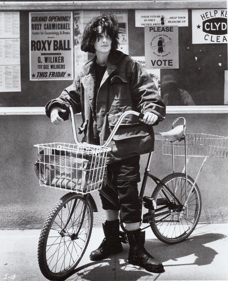Image from Welcome Home, Roxy Carmichael, showing Winona Ryder as Dinky Bosetti, wearing her sloppy oversized all-black clothes, her hair unkempt, with her bike