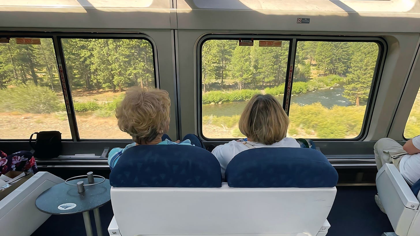 Two women enjoy a view out the window from a train passenger viewing car.