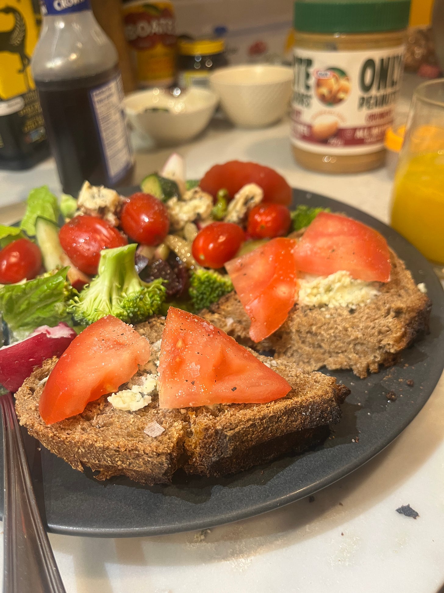 A slice of dark sourdough toast cut in half and topped with crumbled blue cheese and tomato. it's on a colourful plate filled with lots of nice vegetables