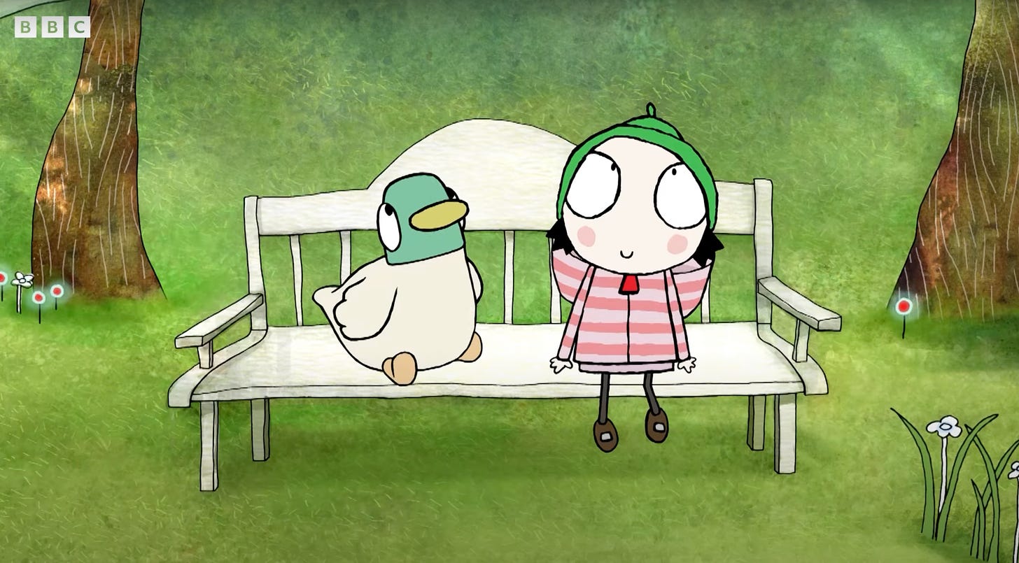 A still from the animated British TV show, Sarah & Duck. Sarah, a little girl with black hair, big eyes, and a green hat sits on a park bench next to her friend, Duck.