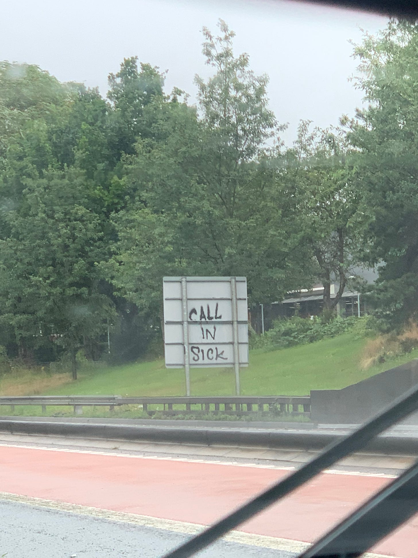 A photo take through a car windscreen on a motorway showing the back of a sign on the opposite side of the road. Graffiti on the sign says: call in sick.