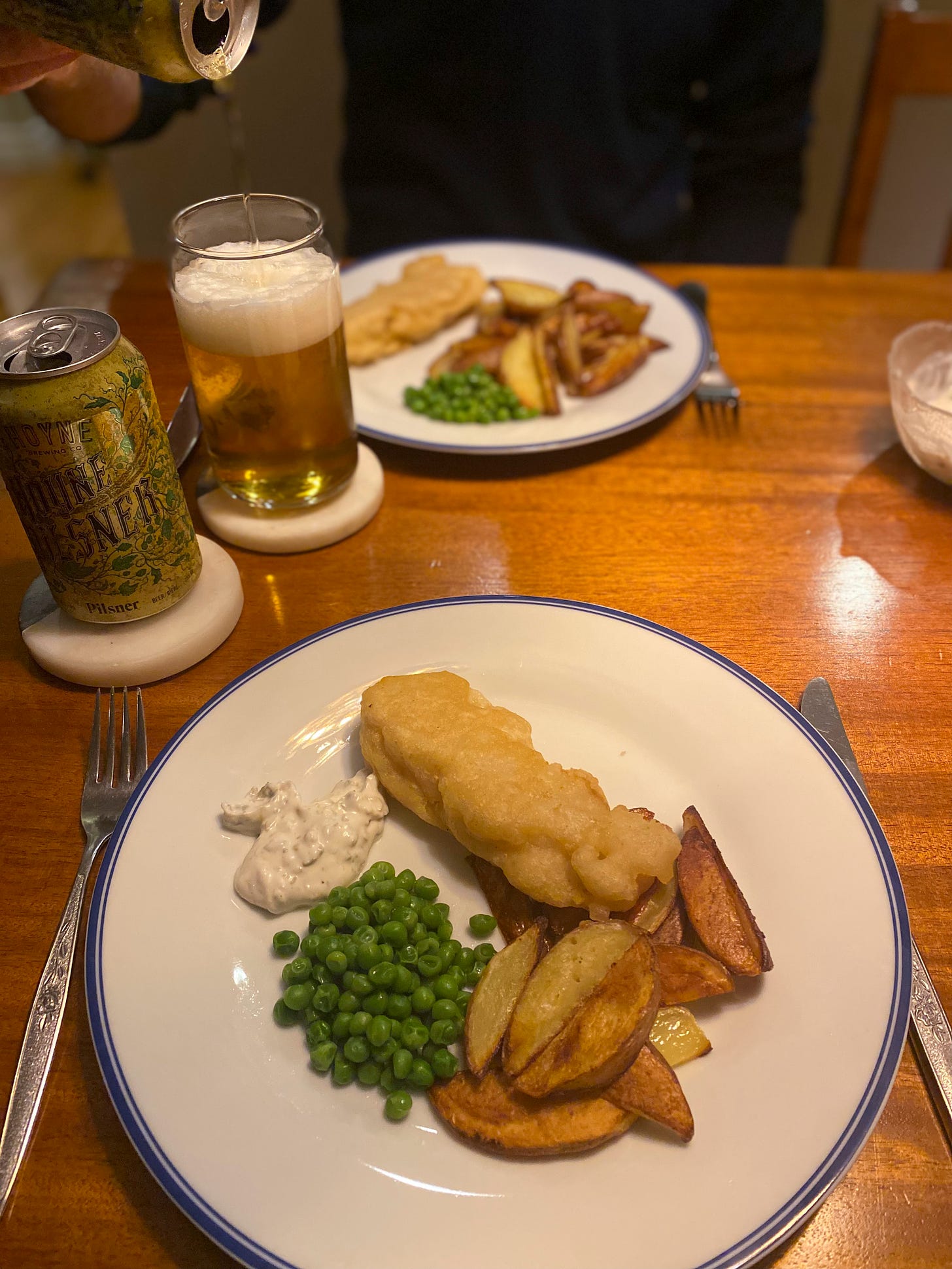 Two plates with pieces of beer-battered cod, wedge fries, tartar sauce, and a small pile of peas. In the background, a can of Hoyne pilsner sits on a coaster, and on another coaster, a second can is being poured into a glass.