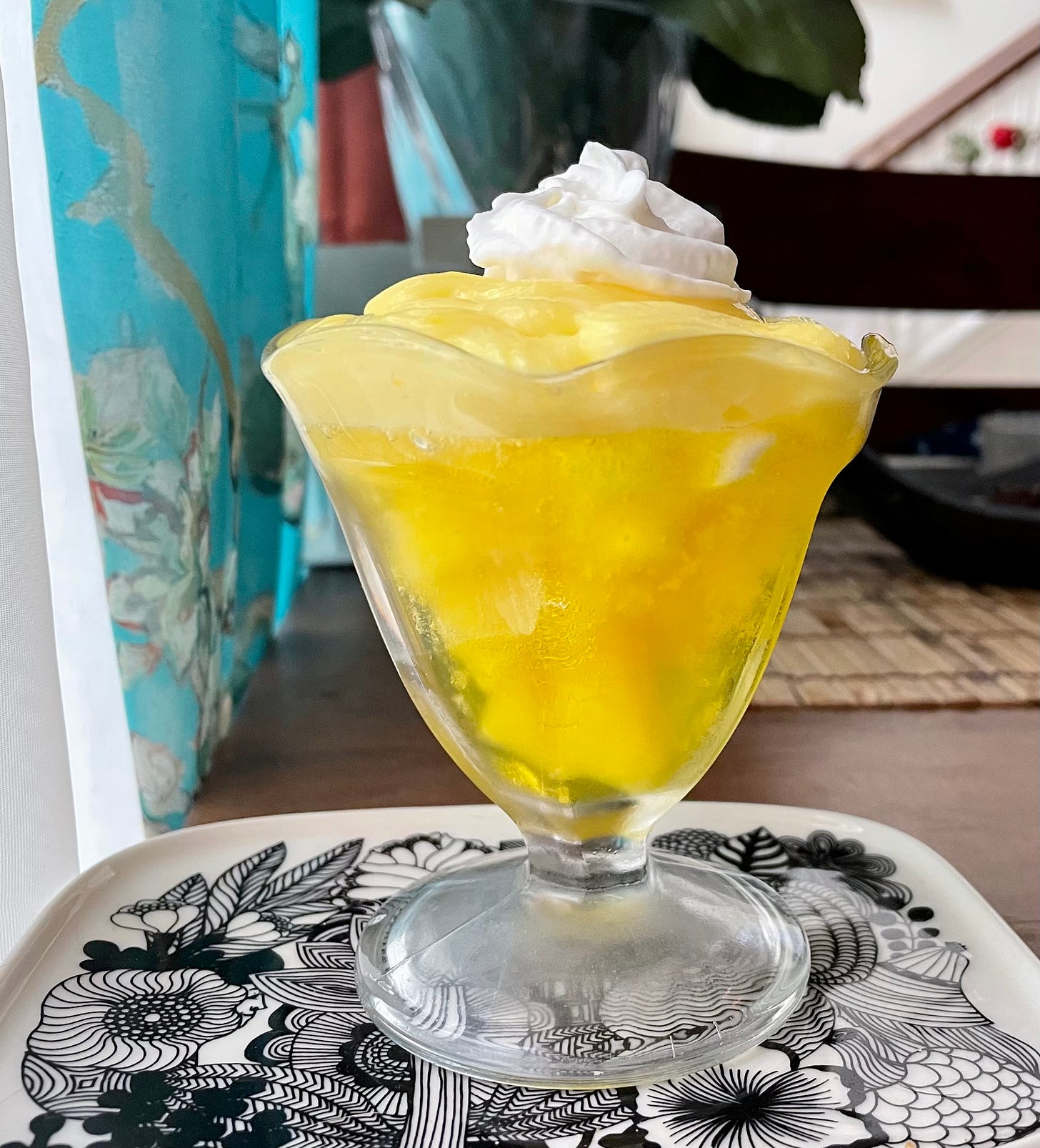 A clear glass sundae dish filled with yellow gelatin studded with hazy yellow fruits and topped with pale lemon pudding, and whipped cream