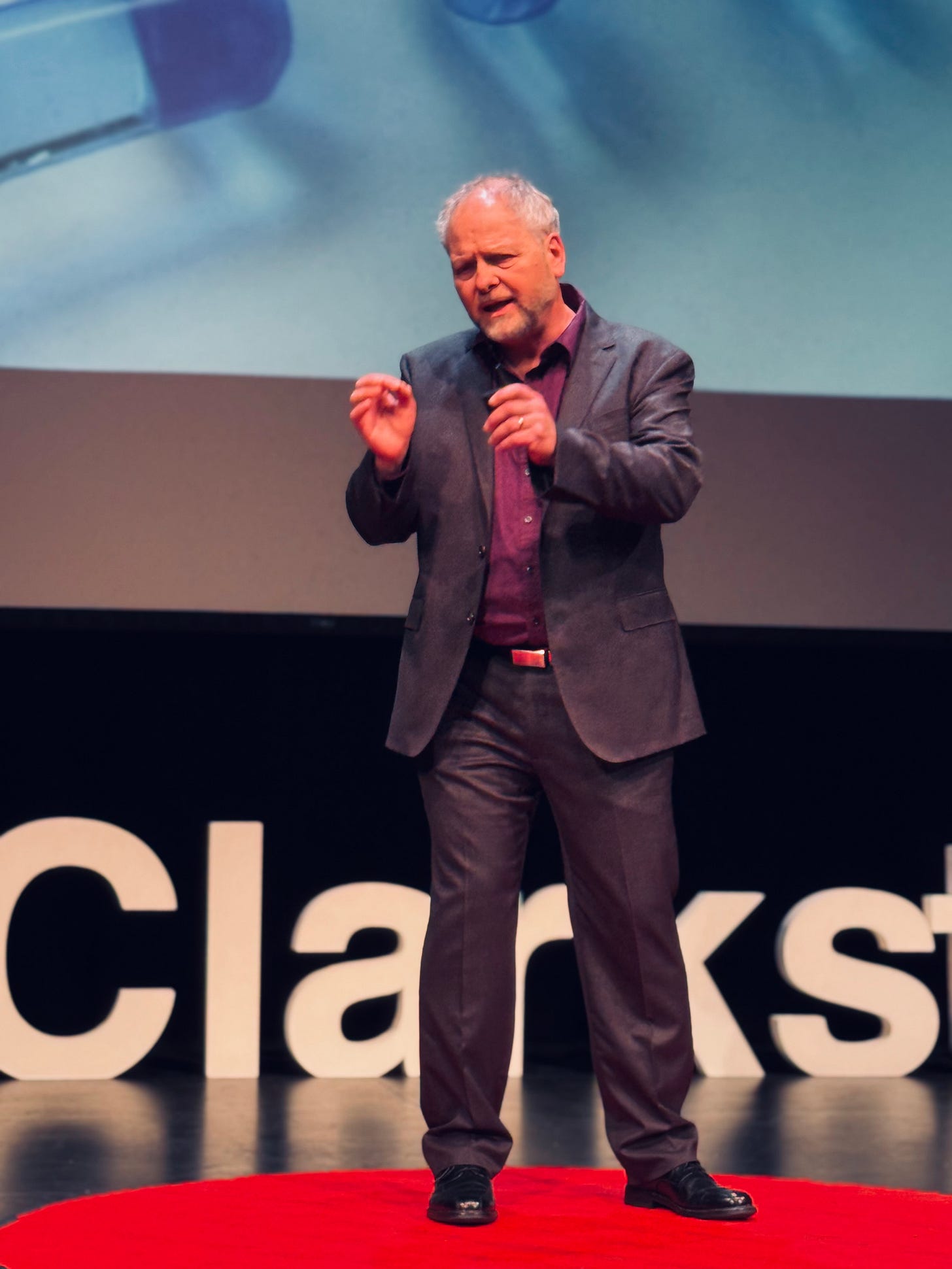 Salvucci speaking on stage in a black suit with a purple shirt. He is looking to his right with his face scrunched in emotion while gesturing. Letters from the TEDX Clarkstown sign are visible behind him. He is standing on the iconic read circle rug.
