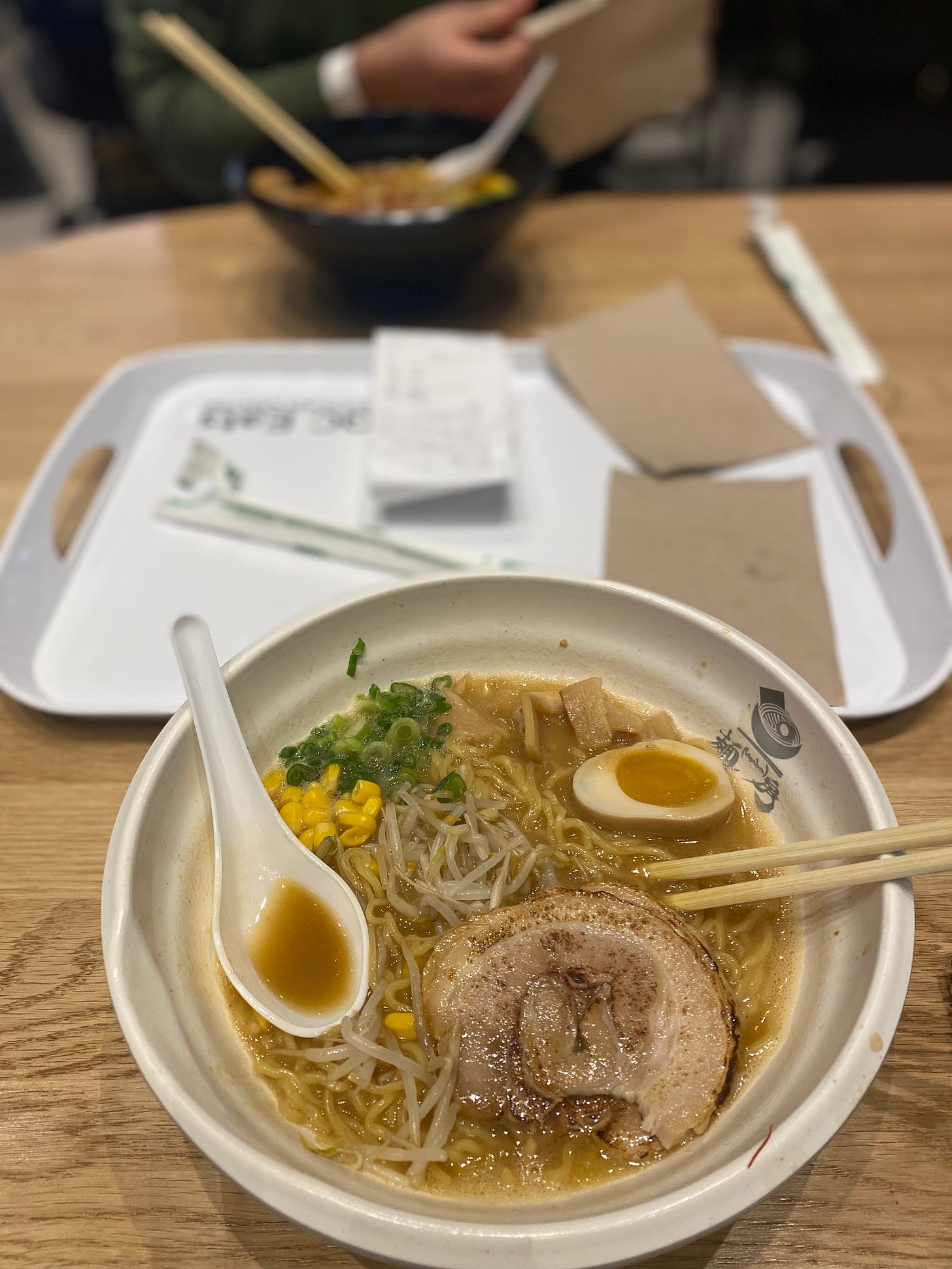 A white bowl of ramen with chashu, corn, bean sprouts, green onions, and half a boiled egg. Jeff's ramen is in a black bowl in the background, and a white tray with brown paper napkins is on the table between them.