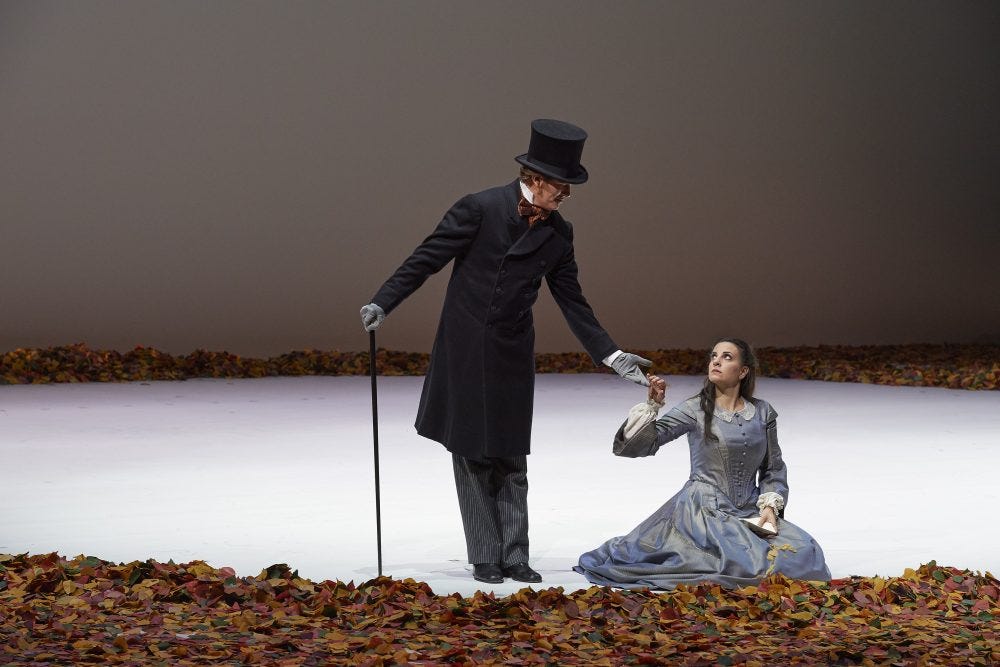 A man in a frock coat and top hat extends his hand to a woman kneeling on the ground and looking up at him. A scene from the opera Eugene Onegin.
