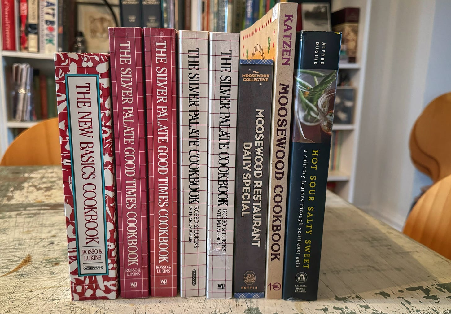 A lineup of cookbooks on a table. From left to right they are: "The New Basics Cookbook," two copies of "The Silver Palate Good Times Cookbook," two copies of "The Silver Palate Cookbook," "Moosewood Restaurant Daily Specials," "The Moosewood Cookbook," and "Hot Sour Salty Sweet: a culinary journey through Southeast Asia"