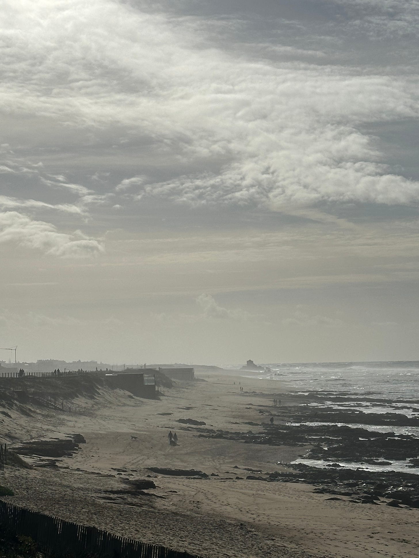 Overcast, grey toned and kind of desolate looking stretch of sandy beach with white thin clouds overhead and low waves foaming to the right. two people walk across in the distance with their backs to the camera. Indescript scatterings of people are seen further out. In the way way way distance is something that might be a church or a lighthouse right against the beach and waves