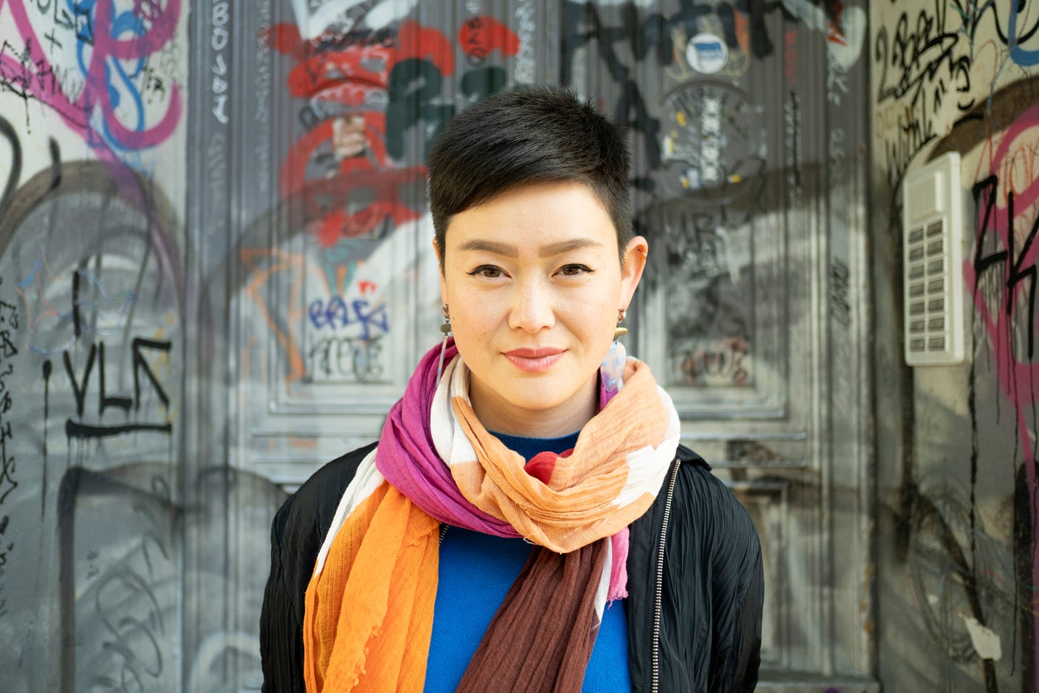 Monika is an Asian woman with short black hair. In this photo, she is standing in front of a grey door covered in graffiti. She is wearing a brightly-coloured scarf on top of a bright blue jumper and black jacket. She is looking straight into the camera, smiling ever so slightly with her mouth closed.