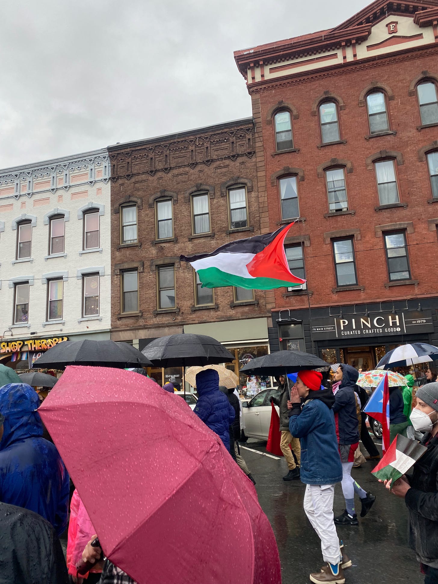 A group of people holding umbrellas march along a main street lined with brick buildings. Someone is waving a Palestinian flag.