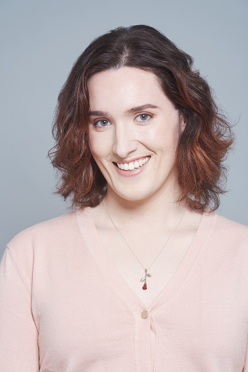 Photo of Abigail Thorn: a smiling young woman with shoulder-length brown hair, wearing a pink top and a rose necklace.  Photo by Ira Giorgetti.