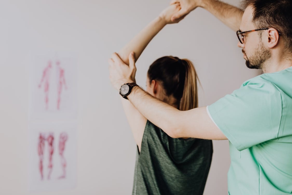 Free Bearded chiropractor in eyeglasses and wristwatch examining arm of anonymous female in casual clothes with raised hand in doctor office in hospital Stock Photo