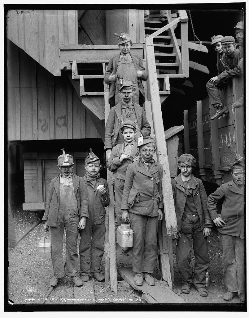 Black-and-white Library of Congress photo showing young ‘breaker boys,’ child laborers at Woodward Coal Mines, Kingston, Pa. c. 1900. The boys are standing, wearing very dirty clothing, with coal dust on their faces, holding metal lunch pails.