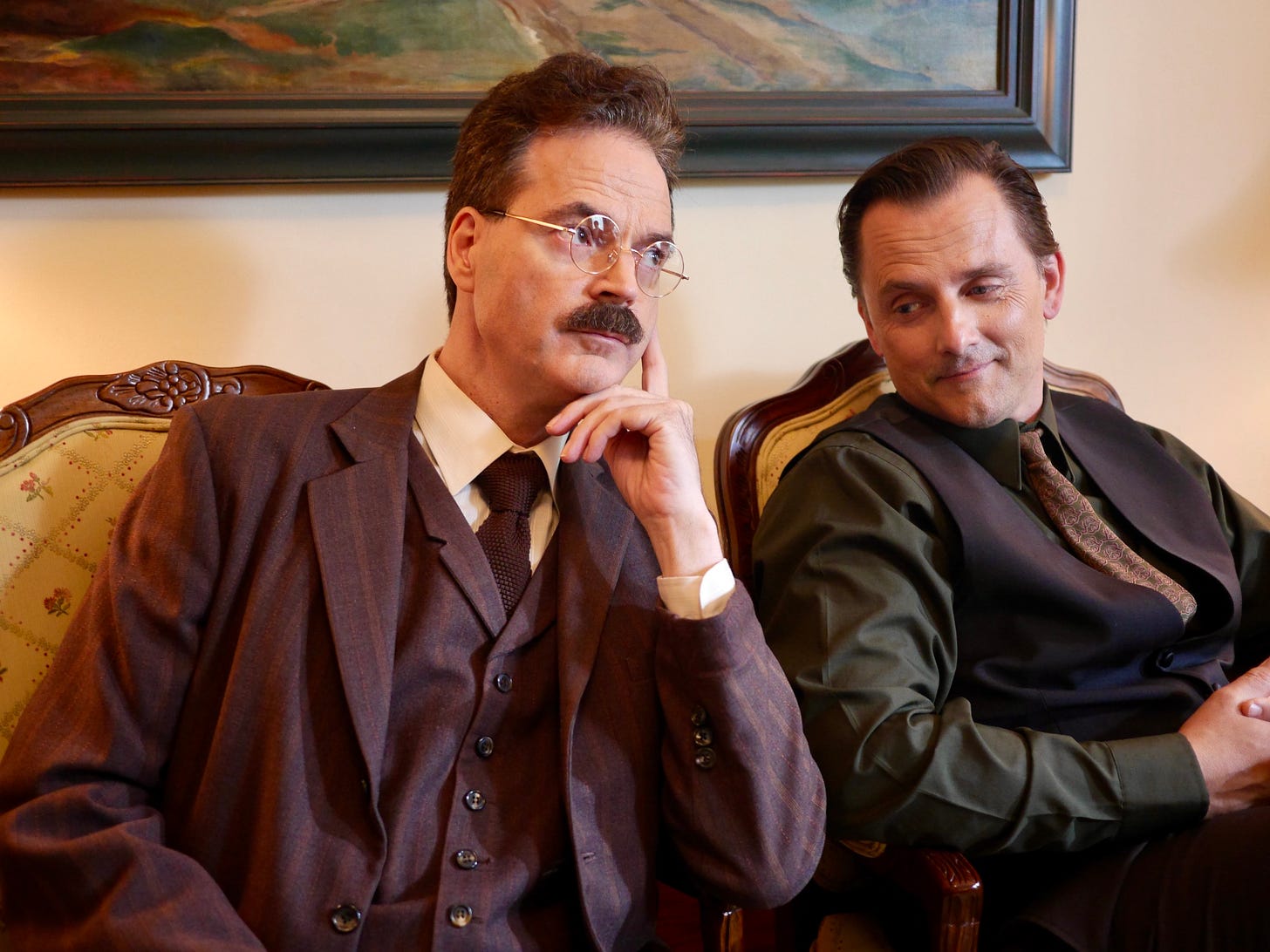 two men sit next to each other under a painting, one in full three-piece suit and spectacles, the other in shirt and waistcoat