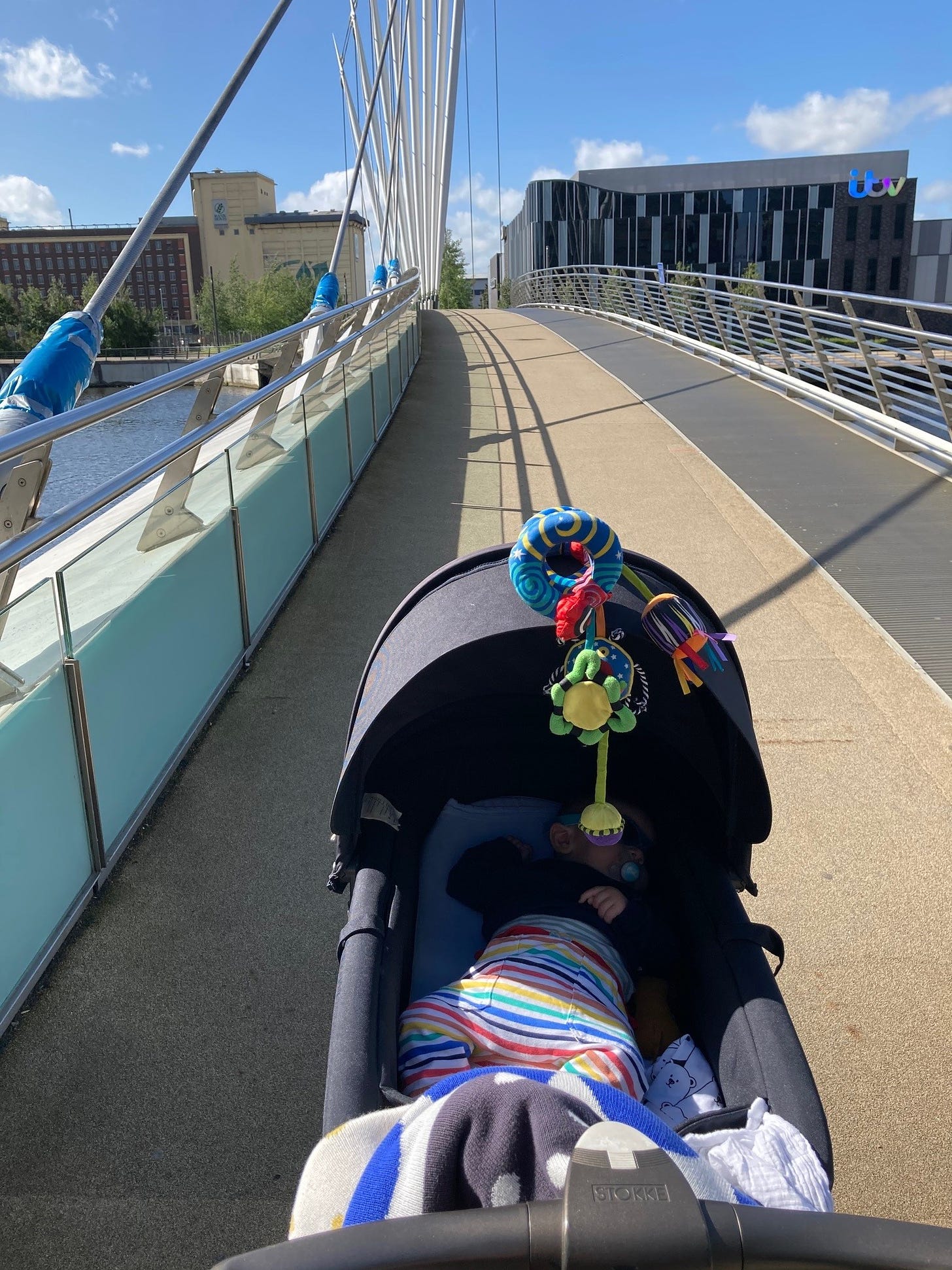 A sunny day. A baby is lay in a pram at the front of the image being pushed over a pedestrian bridge