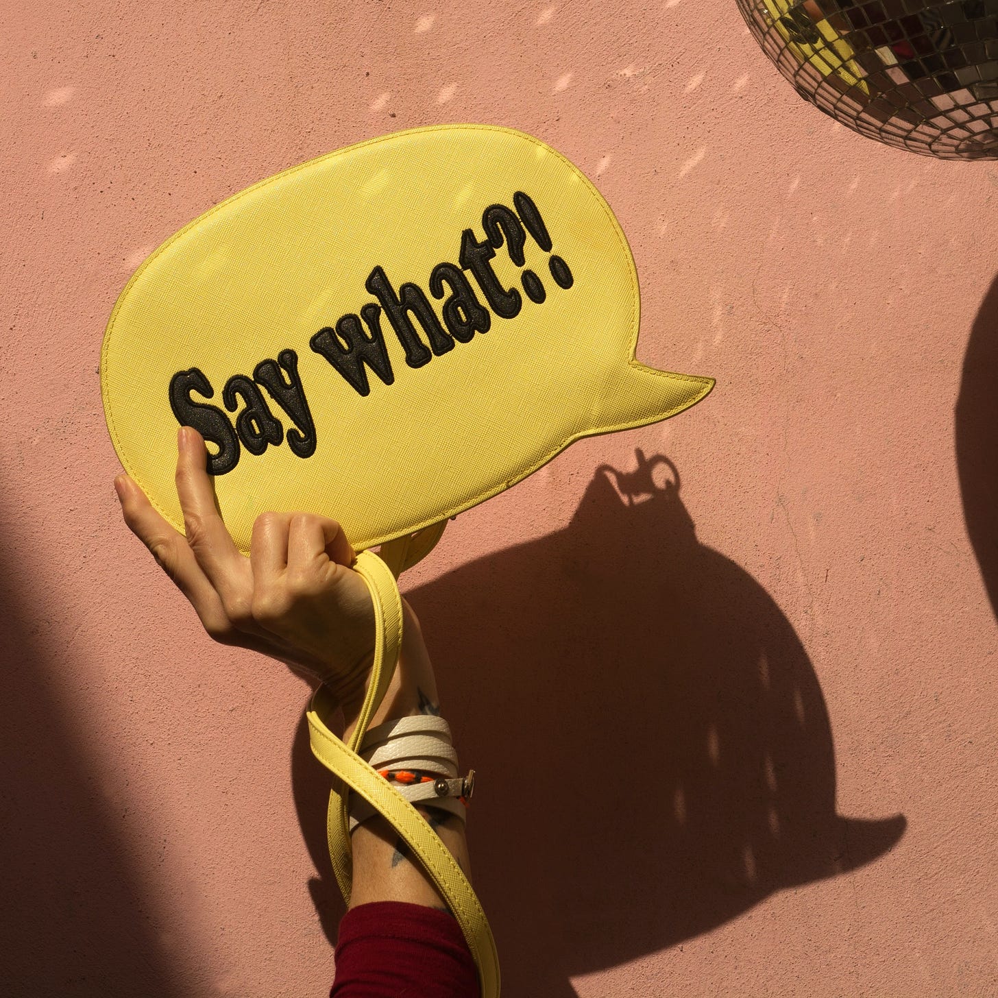 a brown hand holds up a gold bubble with the word "Say what?!" in it over their head