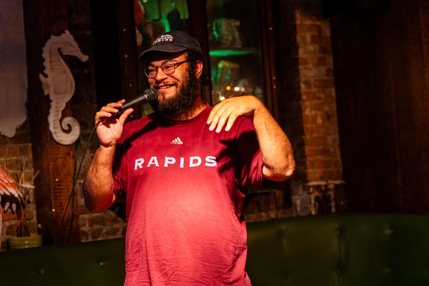 comedian Josh Nasser smiling in glasses and baseball cap with mic at queers n peers variety show