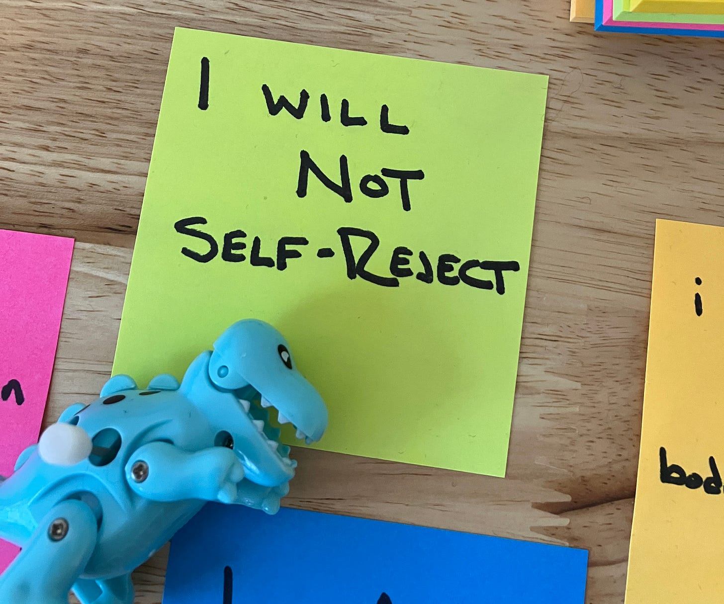 A neon green post-it note on a wood grain background. The post-it reads "I WILL NOT SELF-REJECT" in sharpie. Laid over the post-it note is a light blue wind up dinosaur toy.