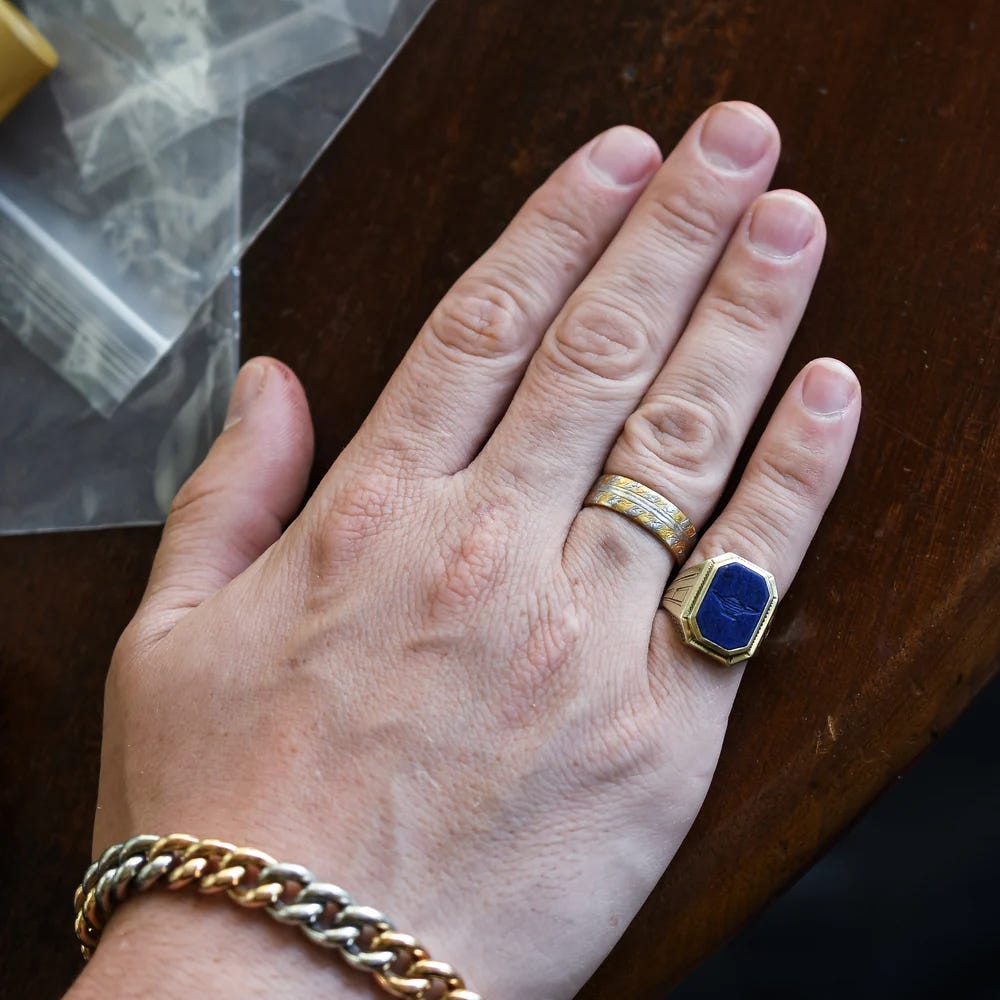 man's hand wearing a gold chain bracelet, gold ring on ring finger and a blue and gold signet ring carved with a genie's lamp on his pinkie