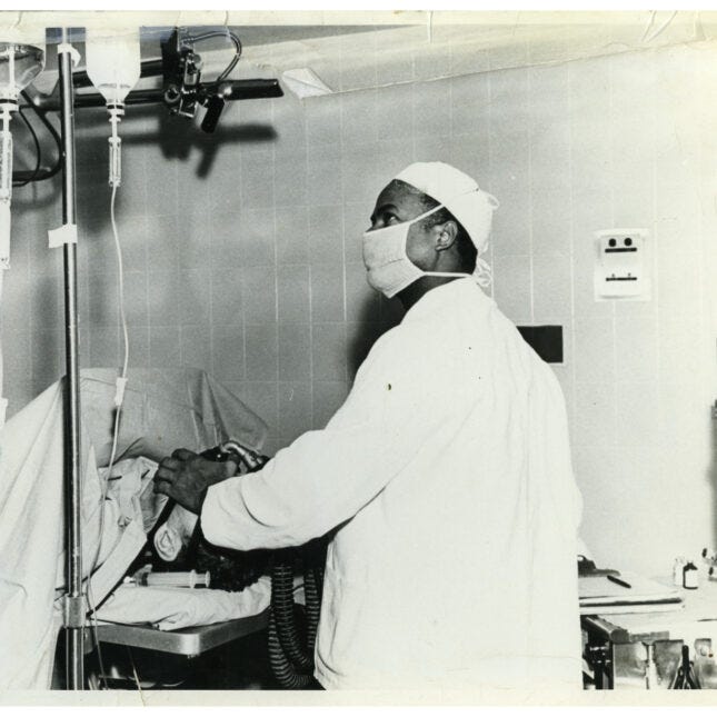 Photograph of Dr. Herman Barnett and looking at some medical equipment while he holds a gas mask over a patient's face. He is wearing a surgical gown, a head covering, and a mask.