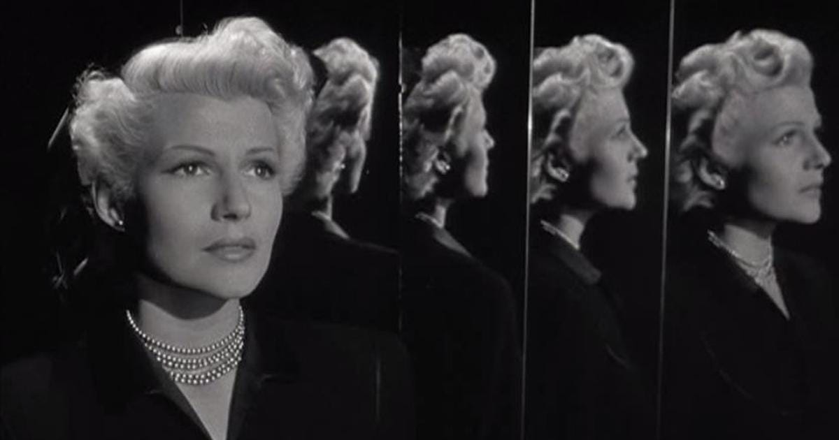 A bleach blond womand in a black dress and pearls. A head and shouldeer shot with her looking out to the right. Her reflection is reflected in multiples mirrors.